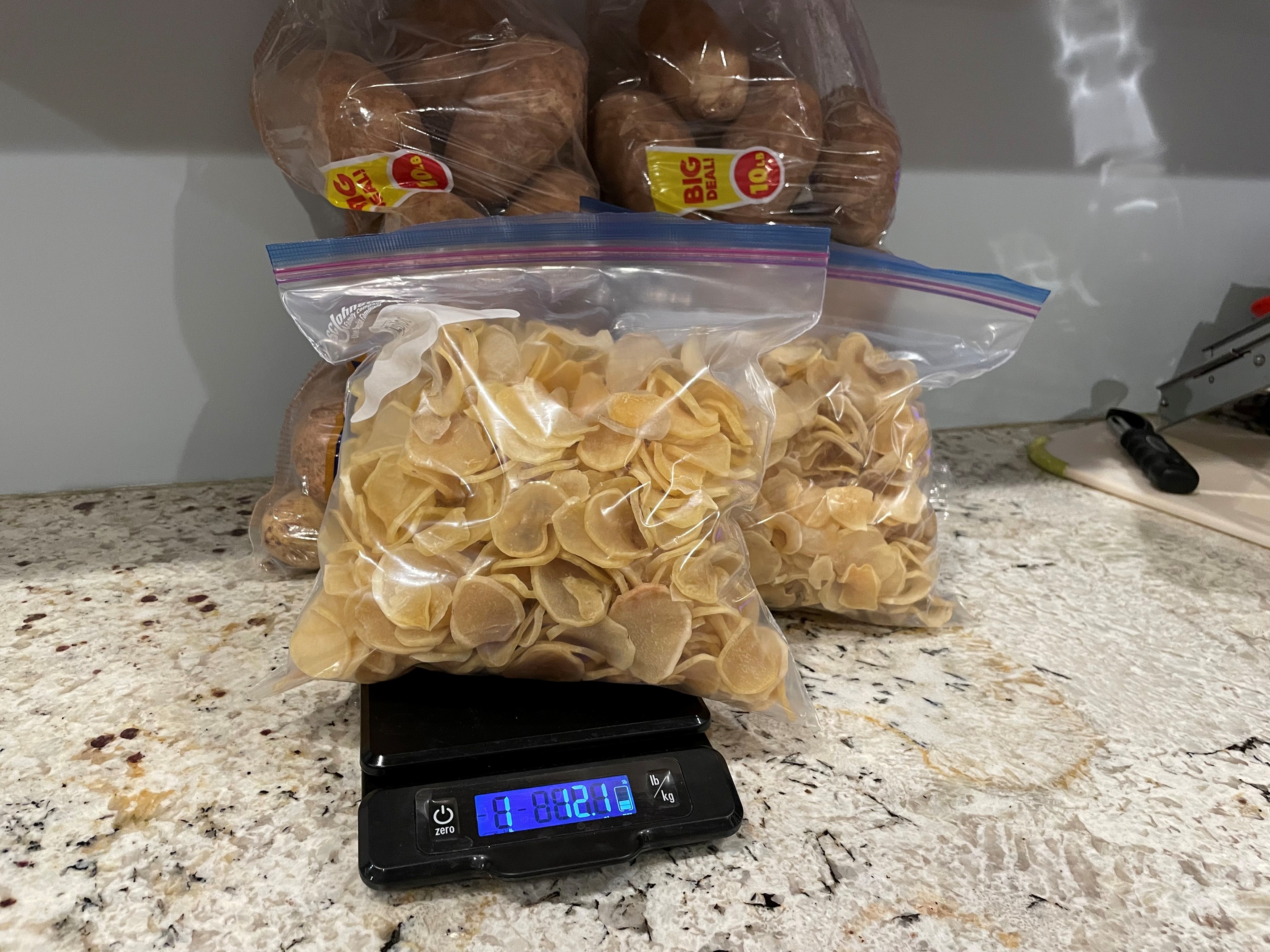Two bags of dried potato slices on a kitchen scale, with bags of fresh russet potatoes behind them.